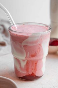 A Hailey Bieber smoothie with a glass straw on a marble counter. The glass is decorated with coconut cream and strawberry jam and the counter has a white cloth napkin and bowl of frozen strawberries in the background.