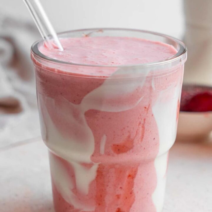 A Hailey Bieber smoothie with a glass straw on a marble counter. The glass is decorated with coconut cream and strawberry jam and the counter has a white cloth napkin and bowl of frozen strawberries in the background.