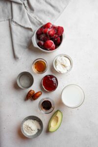 An overhead shot of several bowls in various sizes containing ingredients to make Hailey Bieber smoothie recipe including frozen strawberries, avocado, dates, collagen powder, coconut cream, hyaluronic acid liquid, maple syrup, and vanilla.