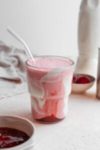 A Hailey Bieber smoothie with a glass straw on a marble counter. The glass is decorated with coconut cream and strawberry jam and the counter has a bowl of jam and bowl of frozen strawberries in the background.