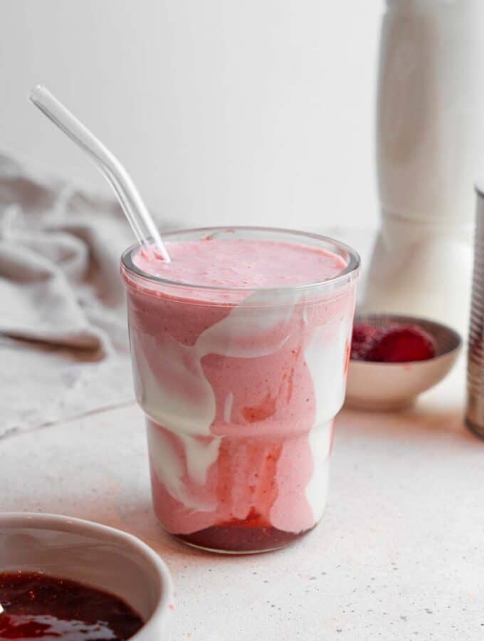A Hailey Bieber smoothie with a glass straw on a marble counter. The glass is decorated with coconut cream and strawberry jam and the counter has a bowl of jam and bowl of frozen strawberries in the background.