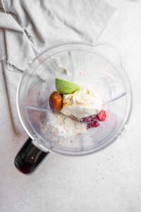 A glass blender containing ingredients to make the Hailey Bieber smoothie including coconut cream, collaged powder, frozen strawberries, avocado, and dates. The blender is on a marble counter with a white cloth napkin next to it.