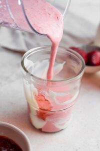 A glass blender pouring the Hailey Bieber smoothie into a glass decorated with coconut cream and jam at the bottom. The glass is on a marble counter with a bowl of strawberries and white cloth napkin in the background.