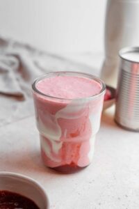 A Hailey Bieber smoothie on a marble counter. The glass is decorated with coconut cream and strawberry jam and the counter has a bowl of jam and frozen strawberries in the background.