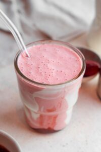 A Hailey Bieber smoothie with a glass straw on a marble counter. The glass is decorated with coconut cream and strawberry jam and the counter has a bowl of frozen strawberries in the background.