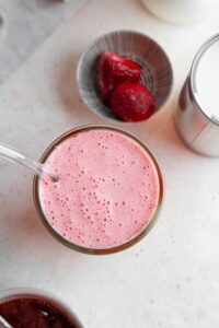 An overhead shot of the Hailey Bieber smoothie in a glass with a glass straw. The smoothie is on a marble counter next to a small bowl of strawberries and sealed metal tin.