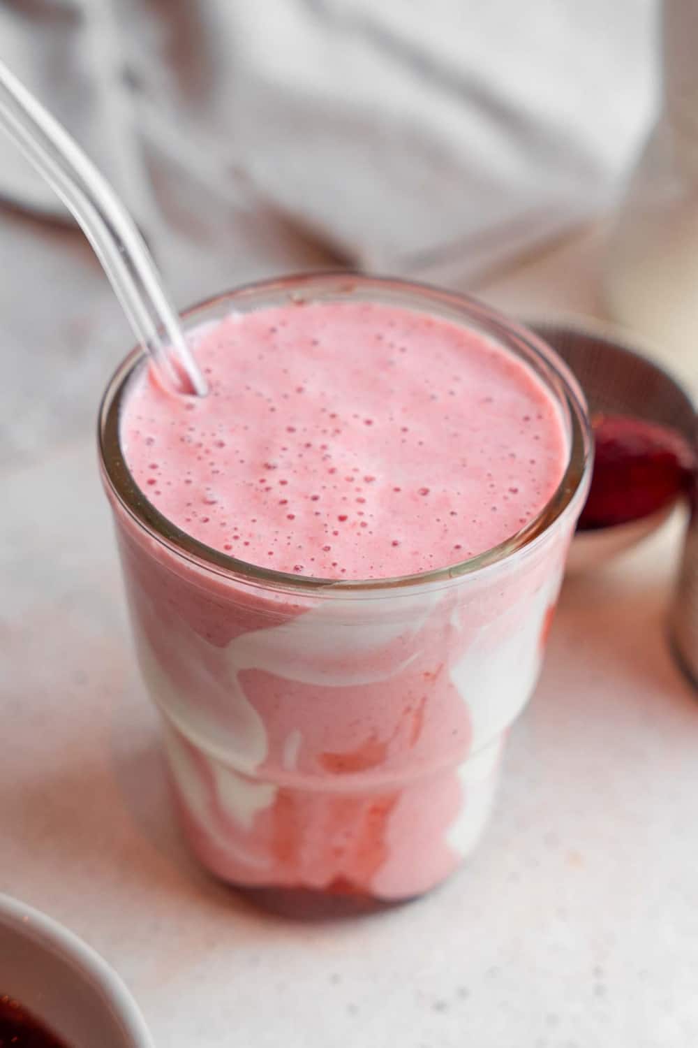 A Hailey Bieber smoothie with a glass straw on a marble counter. The glass is decorated with coconut cream and strawberry jam and the counter has a bowl of frozen strawberries in the background.