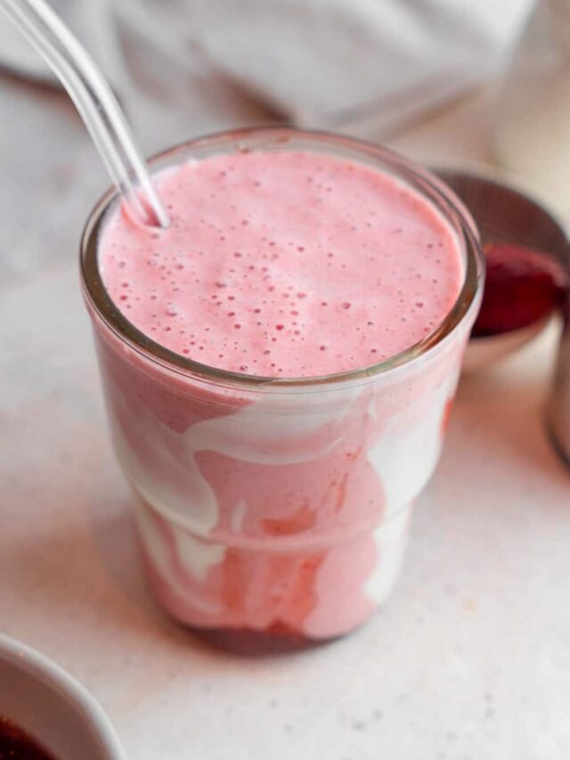 A Hailey Bieber smoothie with a glass straw on a marble counter. The glass is decorated with coconut cream and strawberry jam and the counter has a bowl of frozen strawberries in the background.